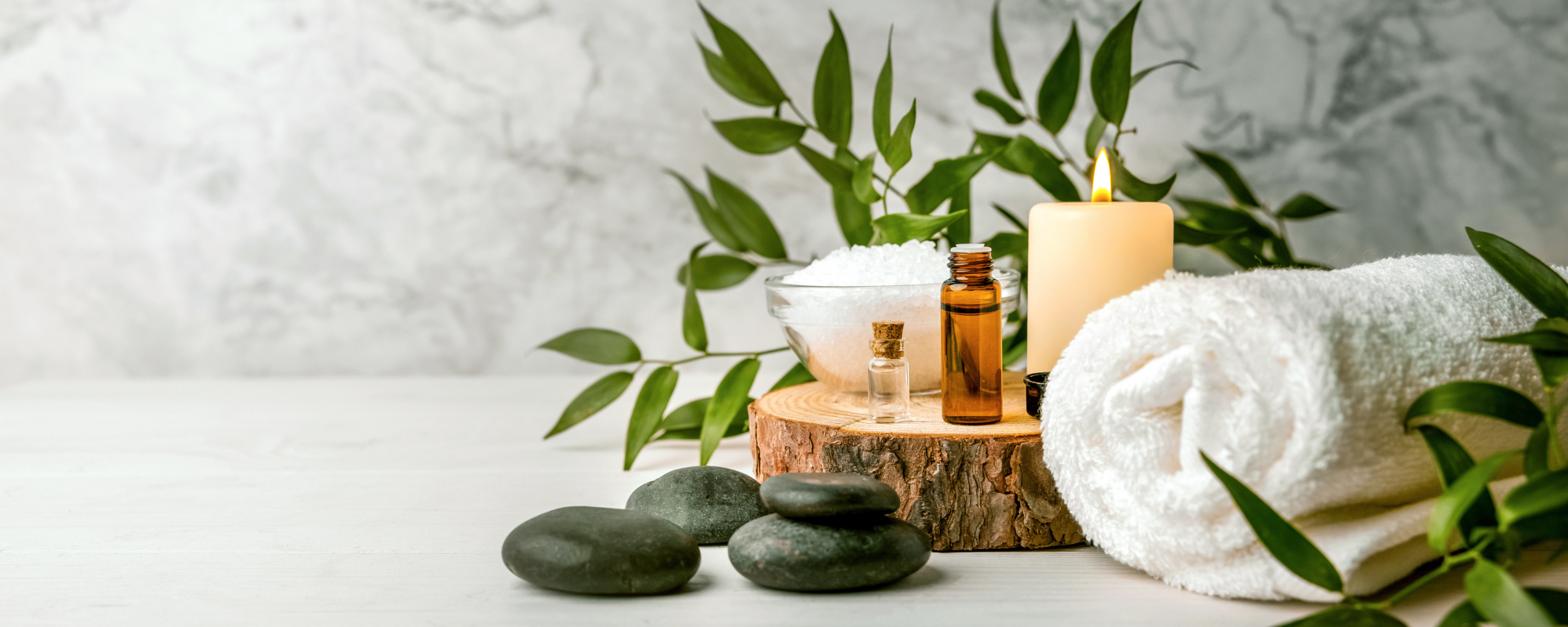 En avkopplande spa-uppläggning med svarta stenar, doftande oljor, havssalt, en tänd ljus och en mjuk handduk på en träskiva, omgiven av gröna växter.